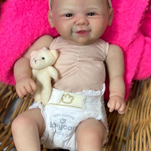 May include: A lifelike baby doll with a smiling expression, wearing a light pink top and a diaper. The doll has a pink bow headband and is holding a small teddy bear. The doll is seated in a woven basket, with a bright pink blanket in the background.