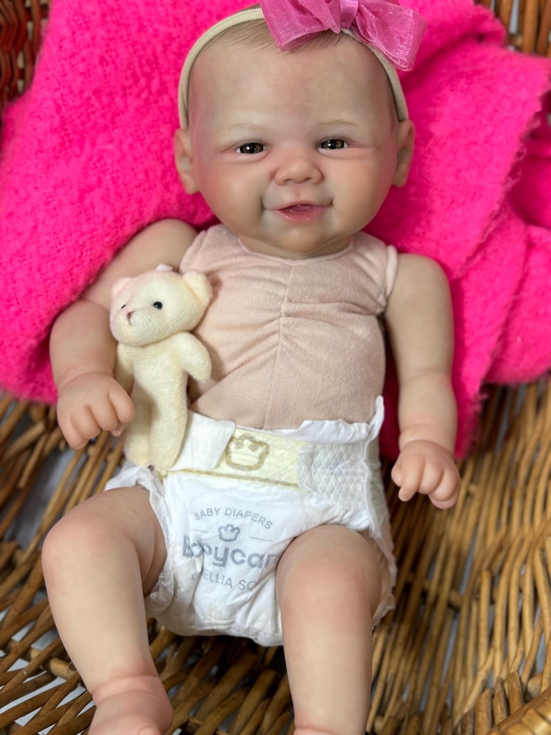 May include: A lifelike baby doll with a smiling expression, wearing a light pink top and a diaper. The doll has a pink bow headband and is holding a small teddy bear. The doll is seated in a woven basket, with a bright pink blanket in the background.