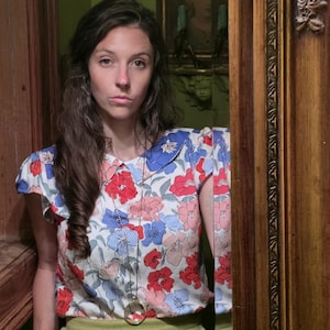 May include: A floral print blouse with red, blue, and pink flowers on a white background. The blouse has a blue collar and short sleeves. The person is wearing a long gold necklace with a circular pendant and green pants.