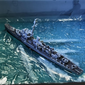 May include: A grey warship model sits in a diorama of ocean water. The model has detailed features like turrets and masts. The water is represented by a textured, blue-green material. The model is a detailed representation of a naval vessel.