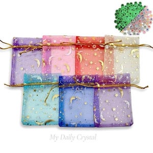 May include: A collection of sheer drawstring pouches in various colors, including purple, pink, and blue. Each pouch is adorned with gold crescent moon and star designs. The pouches are arranged together, with a small pile of star-shaped embellishments in the upper right corner. The text "My Daily Crystal" is at the bottom.