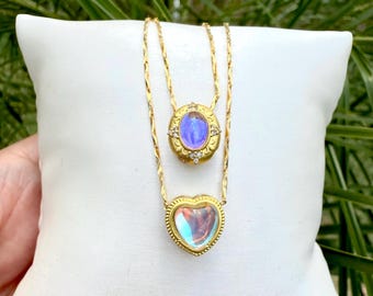 May include: Two gold necklaces with iridescent pendants. The top pendant is oval-shaped with a blue-purple center and small clear stones. The bottom pendant is heart-shaped. Both necklaces are displayed on a white pillow.