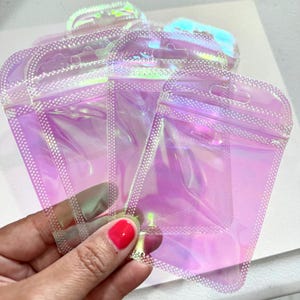 May include: Several iridescent, clear, zip-top pouches are held in a hand. The pouches have a pearlescent sheen, reflecting shades of pink, purple, and green. The edges are sealed with a dotted pattern, and each pouch has a small hole for hanging.