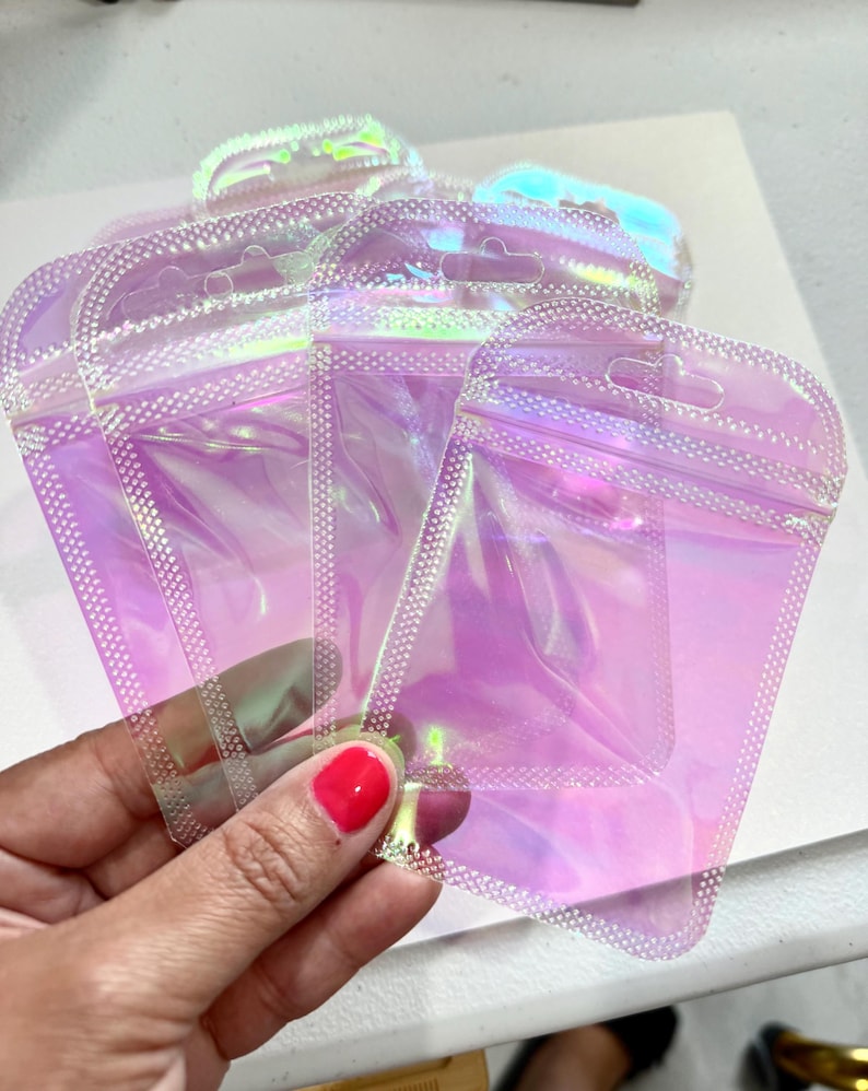 May include: Several iridescent, clear, zip-top pouches are held in a hand. The pouches have a pearlescent sheen, reflecting shades of pink, purple, and green. The edges are sealed with a dotted pattern, and each pouch has a small hole for hanging.