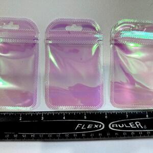 May include: Three iridescent, holographic zip-top pouches with a hang hole at the top. The pouches are rectangular with rounded corners and a white dotted border. A black ruler with inch measurements is at the bottom.