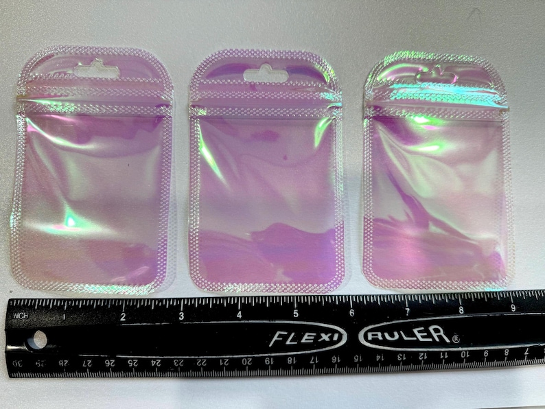 May include: Three iridescent, holographic zip-top pouches with a hang hole at the top. The pouches are rectangular with rounded corners and a white dotted border. A black ruler with inch measurements is at the bottom.