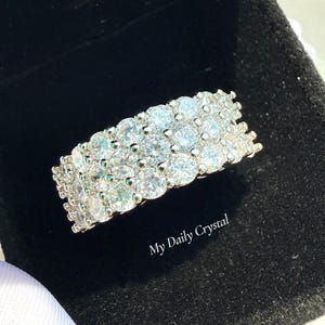 May include: A silver ring with three rows of clear, round-cut gemstones. The ring is displayed on a black velvet surface. The text "My Daily Crystal" is visible on the surface.