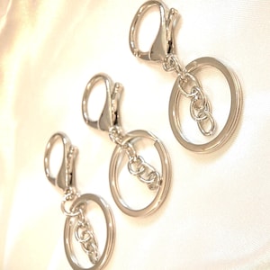 May include: Three silver-colored keychains with a lobster claw clasp, a chain, and a circular key ring. The keychains are displayed on a white, slightly textured surface. The keychains are made of metal and have a shiny finish.