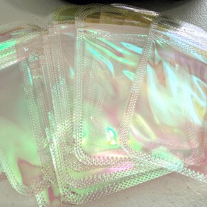 May include: A stack of iridescent, clear plastic pouches with a textured, dotted border. The pouches have a holographic effect, reflecting shades of pink, green, and yellow. The pouches are sealed and appear empty.