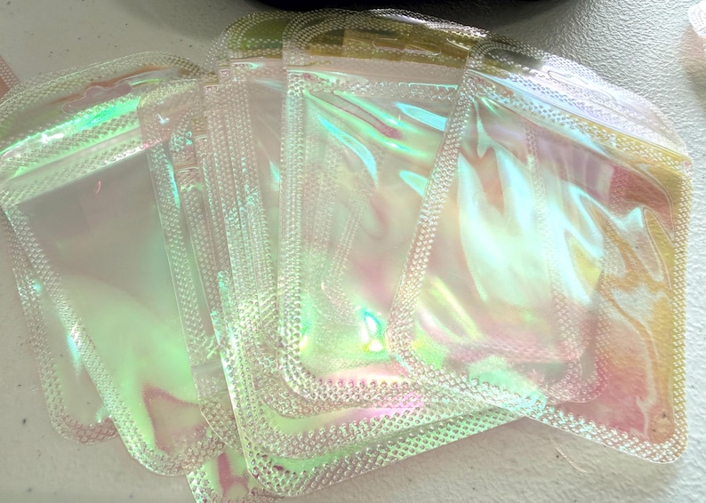 May include: A stack of iridescent, clear plastic pouches with a textured, dotted border. The pouches have a holographic effect, reflecting shades of pink, green, and yellow. The pouches are sealed and appear empty.