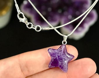 Könnte beinhalten: Eine silberfarbene Schlangenkette mit einem sternförmigen Anhänger aus dunkelviolettem Amethyststein. Der Anhänger ist an einer einfachen Metallöse befestigt, und die Kette verfügt über einen Karabinerverschluss sowie eine kurze Verlängerungskette.