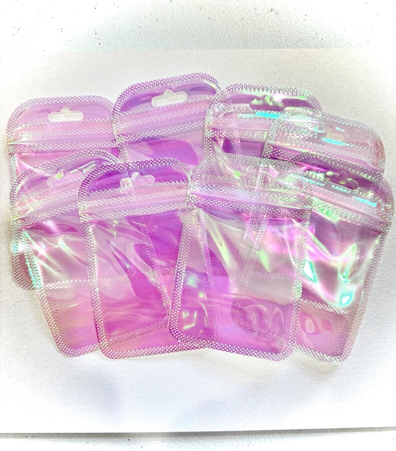 May include: A collection of iridescent, holographic zip-top bags. The bags are a soft pink color with a shimmering, rainbow effect. Each bag has a small hole at the top for hanging. The bags are made of a clear, flexible material.