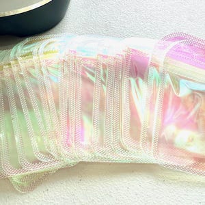 May include: A stack of iridescent, rectangular pouches with a zipper closure. The pouches have a shimmering, rainbow-like effect, reflecting colors like pink, yellow, and green. The pouches are made of a clear, flexible material.