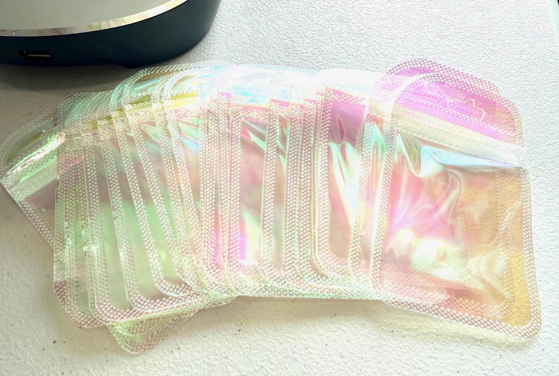 May include: A stack of iridescent, rectangular pouches with a zipper closure. The pouches have a shimmering, rainbow-like effect, reflecting colors like pink, yellow, and green. The pouches are made of a clear, flexible material.