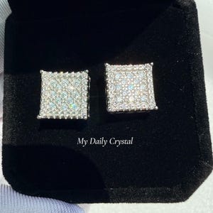 May include: A pair of square-shaped, silver-toned stud earrings, encrusted with numerous small, sparkling crystals. The earrings are displayed in a black velvet jewelry box. The text "My Daily Crystal" is visible.