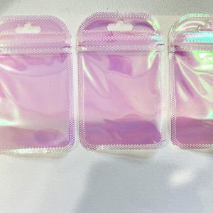 May include: Three iridescent, holographic zip-top pouches with a small hole at the top for hanging. The pouches are a light pink color with a pearlescent sheen. A black ruler with inch measurements is on the left.