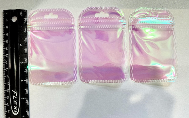 May include: Three iridescent, holographic zip-top pouches with a small hole at the top for hanging. The pouches are a light pink color with a pearlescent sheen. A black ruler with inch measurements is on the left.