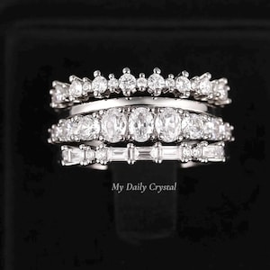 May include: A stack of three silver rings with clear gemstones. The top ring features small, round stones. The middle ring has oval-shaped stones, and the bottom ring has rectangular stones. The rings are displayed against a black background.