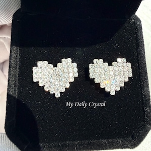 Peut inclure: Boucles d'oreilles en forme de cœur, serties de cristaux étincelants, présentées dans un écrin en velours noir. Le texte "My Daily Crystal" est visible sur la boîte.
