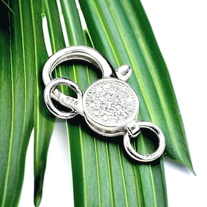 May include: A silver-colored jewelry clasp with a round, gem-studded center and a hinged closure. The clasp has two small rings and is set against a green leaf.