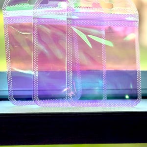 May include: Three iridescent, clear plastic pouches with a zip closure and a small hole at the top for hanging. The pouches have a pearlescent sheen, reflecting colors of pink, purple, and green. The edges are lined with small, white dots.