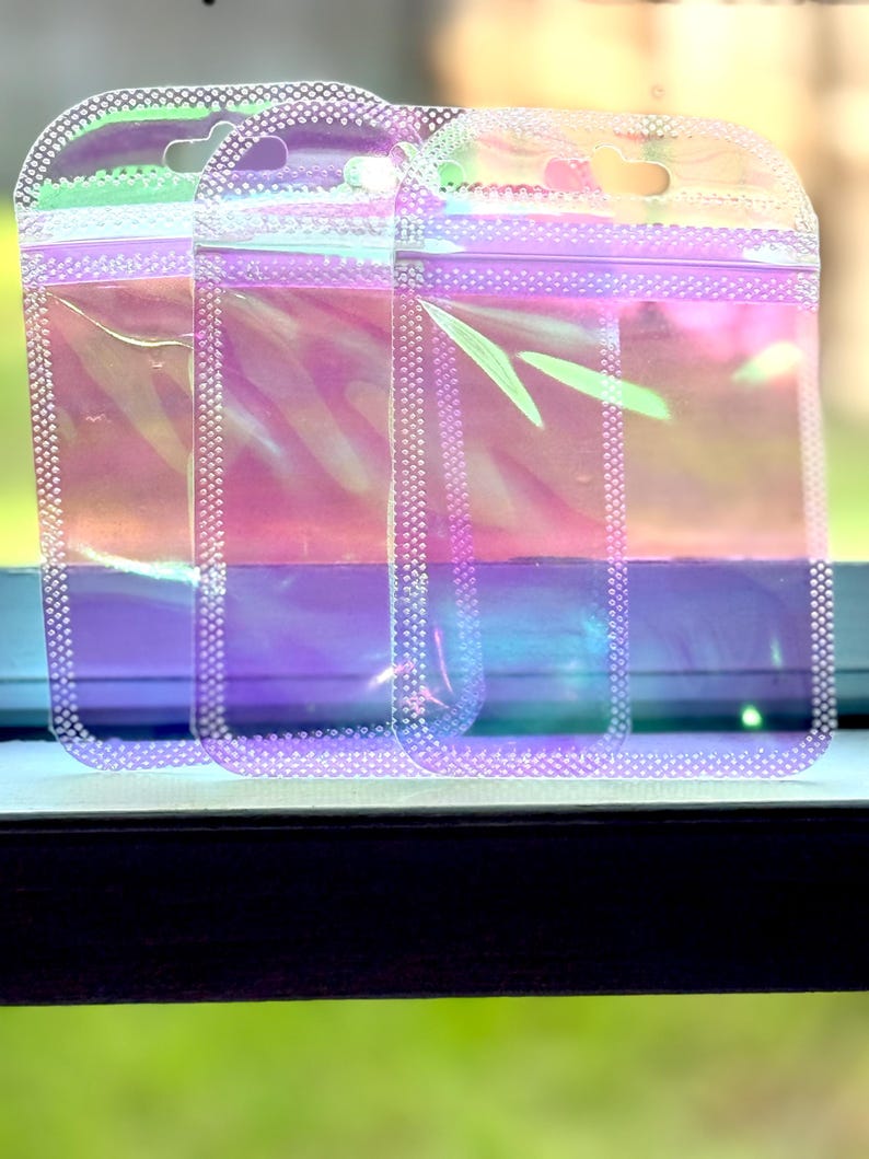 May include: Three iridescent, clear plastic pouches with a zip closure and a small hole at the top for hanging. The pouches have a pearlescent sheen, reflecting colors of pink, purple, and green. The edges are lined with small, white dots.