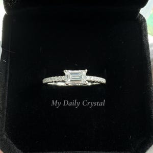 May include: A diamond engagement ring with an emerald-cut center stone, set in a pave band. The ring is displayed in a black ring box. The text "My Daily Crystal" is visible at the bottom of the box.