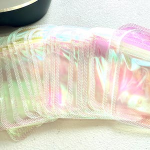 May include: A stack of iridescent, rectangular pouches with a zipper closure. The pouches have a pearlescent sheen, reflecting colors of pink, yellow, and green. The pouches are stacked on a white surface.
