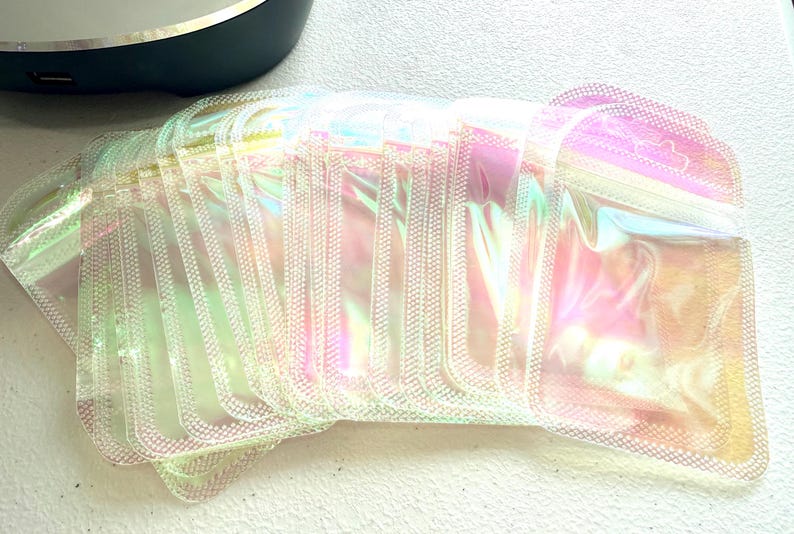 May include: A stack of iridescent, rectangular pouches with a zipper closure. The pouches have a pearlescent sheen, reflecting colors of pink, yellow, and green. The pouches are stacked on a white surface.