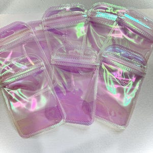 May include: Several iridescent, clear plastic pouches with a zip-top closure are arranged on a white surface. The pouches have a pearlescent sheen, reflecting shades of pink, purple, and green. The edges are outlined with a dotted pattern.