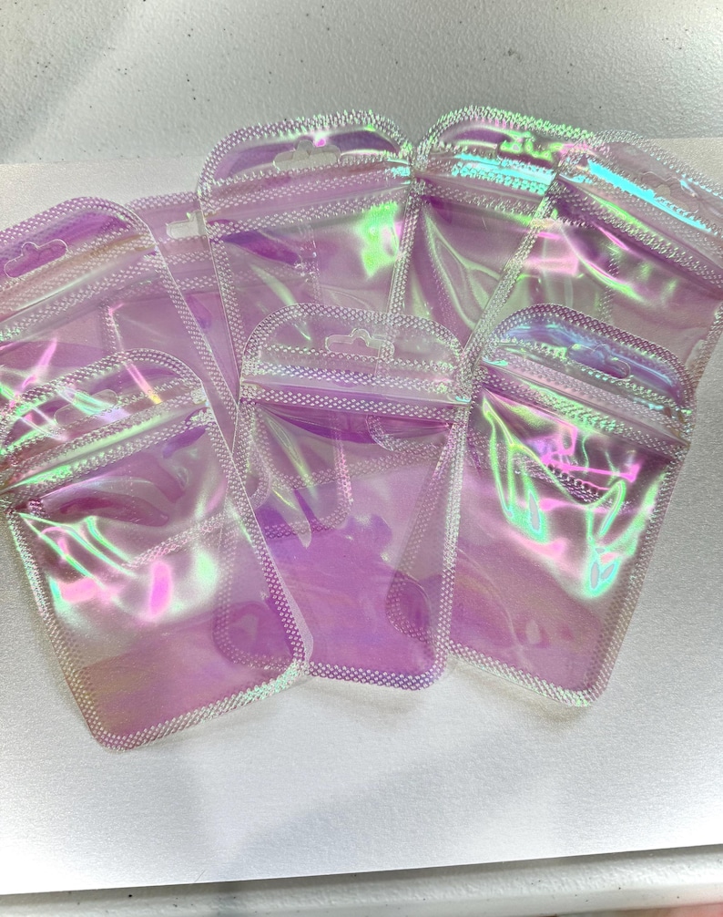 May include: Several iridescent, clear plastic pouches with a zip-top closure are arranged on a white surface. The pouches have a pearlescent sheen, reflecting shades of pink, purple, and green. The edges are outlined with a dotted pattern.