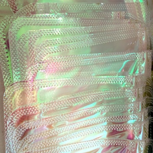 May include: A stack of iridescent, clear plastic pouches. Each pouch has a rounded corner and a textured, dotted border. The pouches reflect a range of colors, including pink, green, and yellow, creating a shimmering effect.