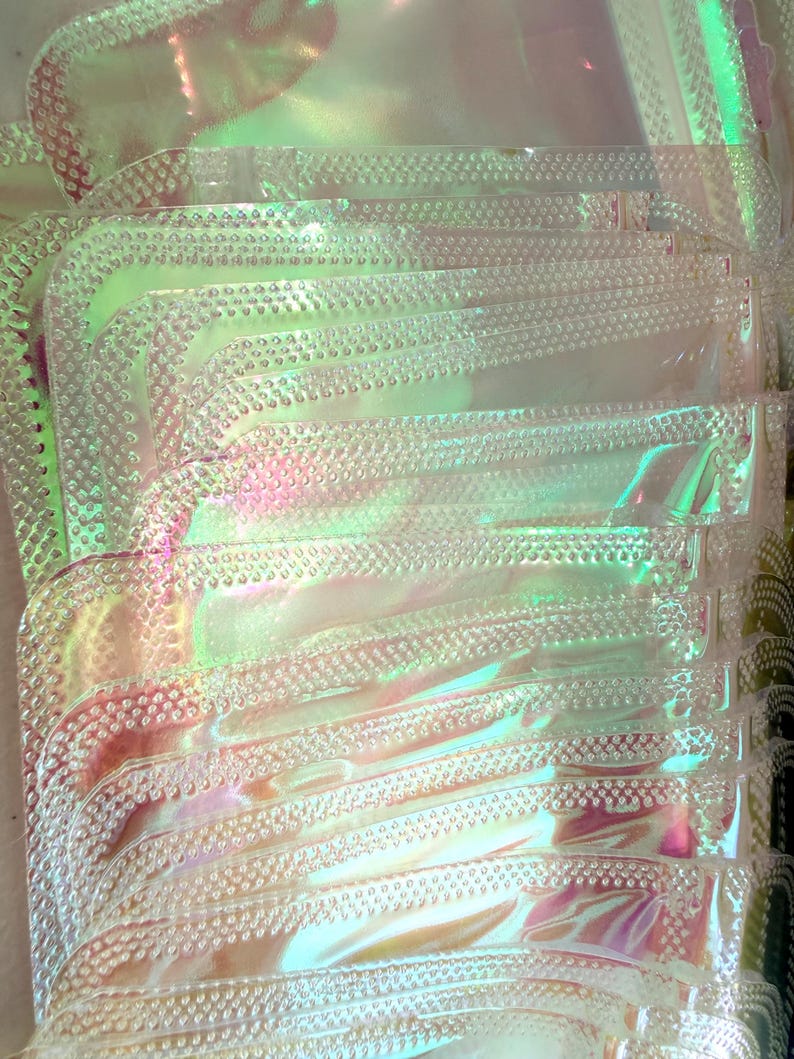 May include: A stack of iridescent, clear plastic pouches. Each pouch has a rounded corner and a textured, dotted border. The pouches reflect a range of colors, including pink, green, and yellow, creating a shimmering effect.