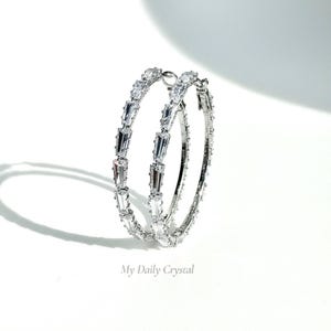 May include: A pair of silver hoop earrings with clear, rectangular crystals set along the outer edge. The earrings are displayed on a white background with the text "My Daily Crystal".