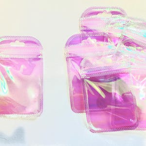 May include: Several iridescent pink, clear, and purple pouches with a zip closure and a small hole for hanging. The pouches are made of a shiny, transparent material. The pouches are empty and ready to be filled.