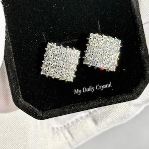 May include: Square-shaped stud earrings with a dazzling array of small, clear crystals. The earrings are set in a silver-toned metal and presented in a black velvet jewelry box. The text "My Daily Crystal" is visible.