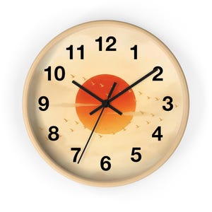 Könnte beinhalten: Runde Wanduhr mit hellem Holzrahmen. Das Zifferblatt zeigt ein Sonnenuntergangsdesign mit einer großen orangefarbenen Sonne, kleinen Vögeln und schwarzen Zeigern. Die Zahlen sind schwarz.