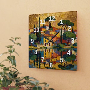 Op de afbeelding: Vierkante wandklok met een kleurrijk landschapsschilderij van een dorp dat weerspiegeld wordt in het water. De klok heeft witte cijfers en zwarte wijzers. Het kunstwerk bevat bomen, gebouwen en een gouden achtergrond.