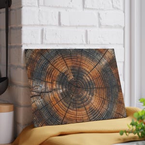 May include: A rectangular wooden cutting board with a detailed cross-section of a tree trunk. The board features concentric rings in shades of brown and black, showcasing the wood's natural grain and texture. The board is resting on a yellow cloth.