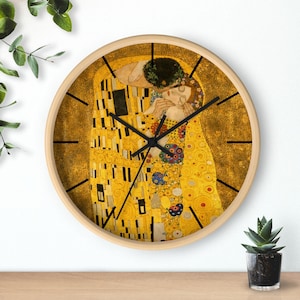 Puede incluir: Reloj de pared redondo con la obra "El Beso" de Gustav Klimt. El reloj tiene un marco de madera clara, manecillas negras y un fondo dorado. La obra representa a una pareja abrazándose, con patrones intrincados y detalles de pan de oro.