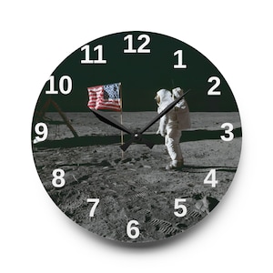 May include: Round wall clock featuring an image of an astronaut on the moon with the American flag. The clock face is black with white numerals and black clock hands. A unique decorative piece.