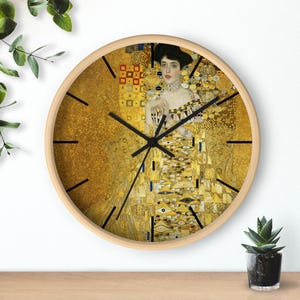 Puede incluir: Reloj de pared redondo con marco de madera clara. La esfera del reloj presenta una reproducción de "Retrato de Adele Bloch-Bauer I" de Gustav Klimt en tonos dorados, amarillos y negros. Agujas y marcadores negros.