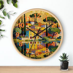 Op de afbeelding: Ronde wandklok met een licht houten frame. De wijzerplaat toont een kleurrijk schilderij van een dorpsgezicht met oranje cijfers. De wijzers zijn zwart en het kunstwerk weerspiegelt in het water eronder.