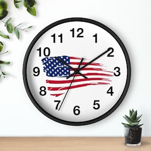 Może przedstawiać: Okrągły zegar ścienny z czarną ramą i białą tarczą. Tarcza zegara przedstawia amerykańską flagę w kolorach czerwonym, białym i niebieskim. Czarne wskazówki wskazują godzinę. Zegar znajduje się na drewnianej powierzchni.