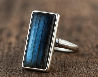 May include: A silver ring with a rectangular, dark blue gemstone. The gemstone has a vertical striped pattern with hints of blue and black. The ring is set on a wooden surface.
