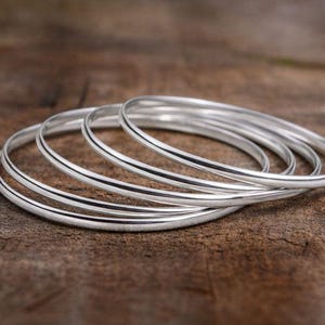 Può includere: Una collezione di braccialetti d'argento, ciascuno con un design liscio e arrotondato. I braccialetti sono impilati su una superficie di legno marrone strutturata. La luce si riflette sul metallo lucido.