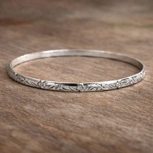 May include: A silver bangle bracelet with a floral pattern. The round bracelet has a repeating design of flowers and leaves. The metal has a polished finish, and the bracelet is shown on a wooden surface.