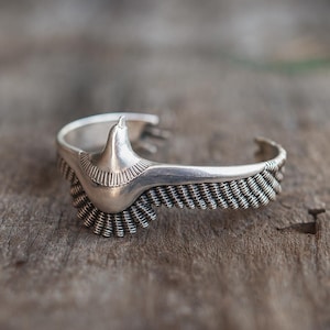 May include: A silver cuff bracelet with an eagle design. The bracelet features a detailed eagle head and wing design. The wings are crafted with a woven texture. The bracelet is set against a wooden surface.
