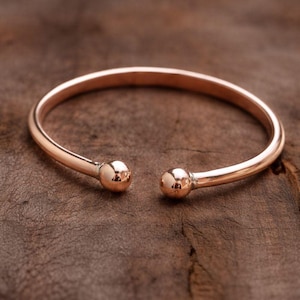 May include: A rose gold cuff bracelet with a simple, open design. The bracelet features a smooth, rounded band that tapers to two small, spherical ends. The jewelry is displayed on a rustic, wooden surface.