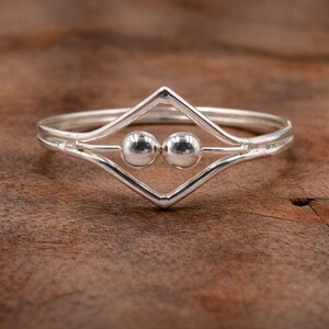 May include: A silver ring with a geometric design. The ring features a diamond-shaped center with two small, round beads. The band is thin and smooth, with a double-band design. The ring is displayed on a wooden surface.